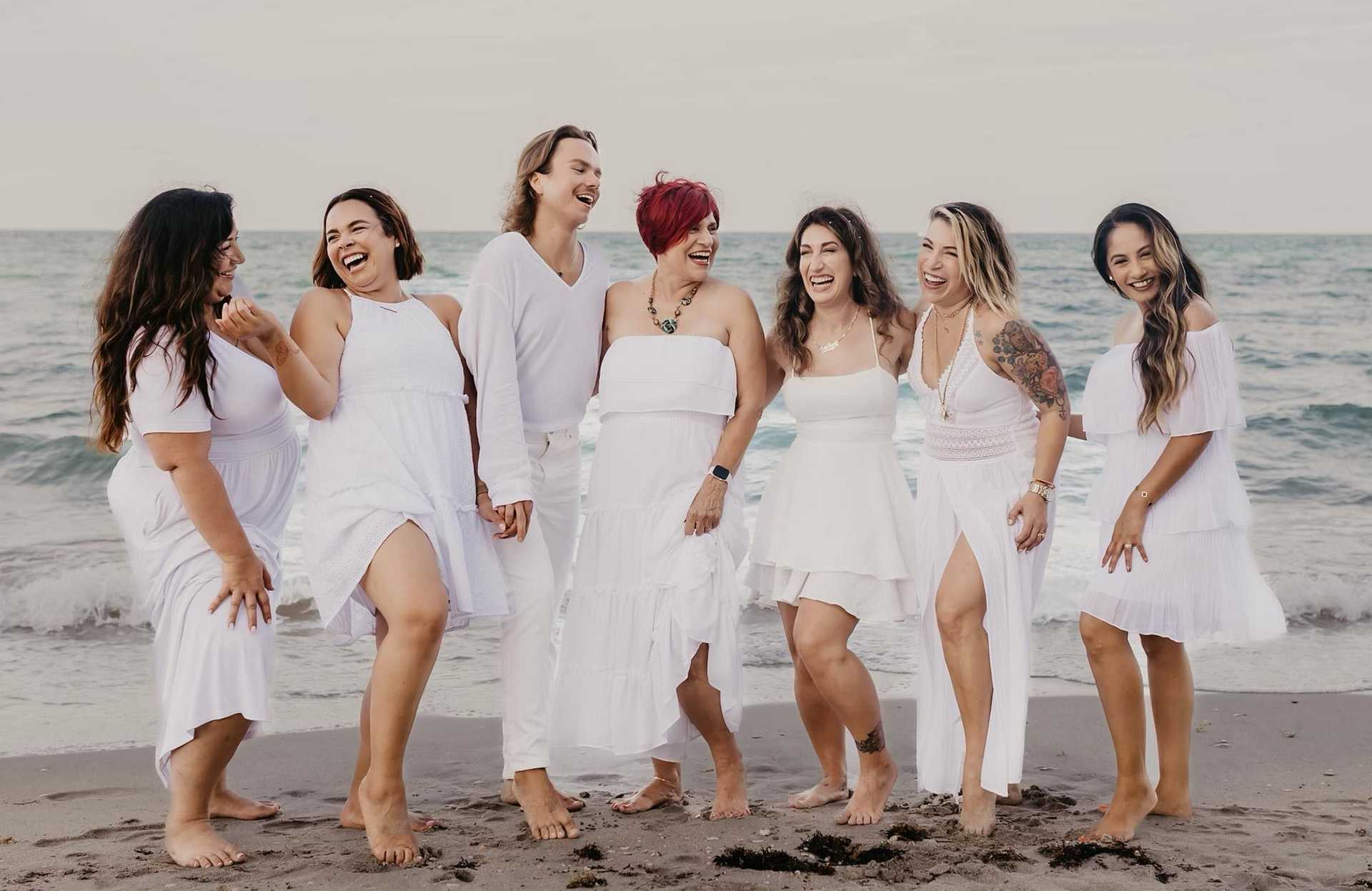 Group of people in white outfits laughing together on a beach.